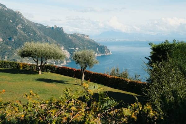 7-Day Amalfi Coast Itinerary: Planned Around How It Feels To Be There 6 A Serene Coastal Landscape Featuring Two Olive Trees On A Lush Green Lawn. A Hedge Overlooks A Deep Blue Sea Framed By Distant, Rugged Mountains.