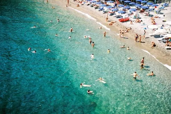 Aerial view of a beach with turquoise waters on the Amalfi Coast. People swim and relax under blue umbrellas on the sandy shore, capturing a lively, sunny atmosphere.